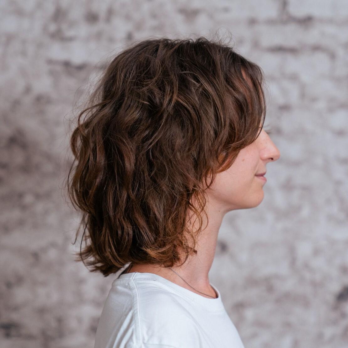 Profilaufnahme einer Person mit schulterlangem Mullet. Die Frisur kombiniert weiche Locken mit gestufter Nackenl&auml;nge.